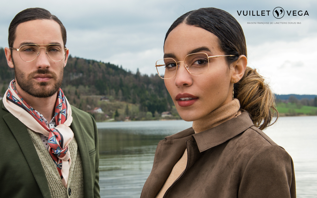 A man and a woman stylishly dressed in eyeglasses stand before a scenic lake with hills in the background.